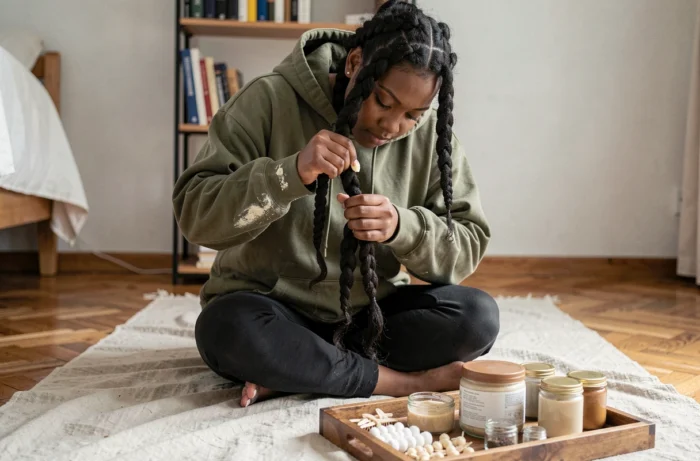Jeune femme afro tressant ses cheveux crépus en twists avec du beurre de karité, assise sur le sol de sa chambre entourée de produits capillaires naturels